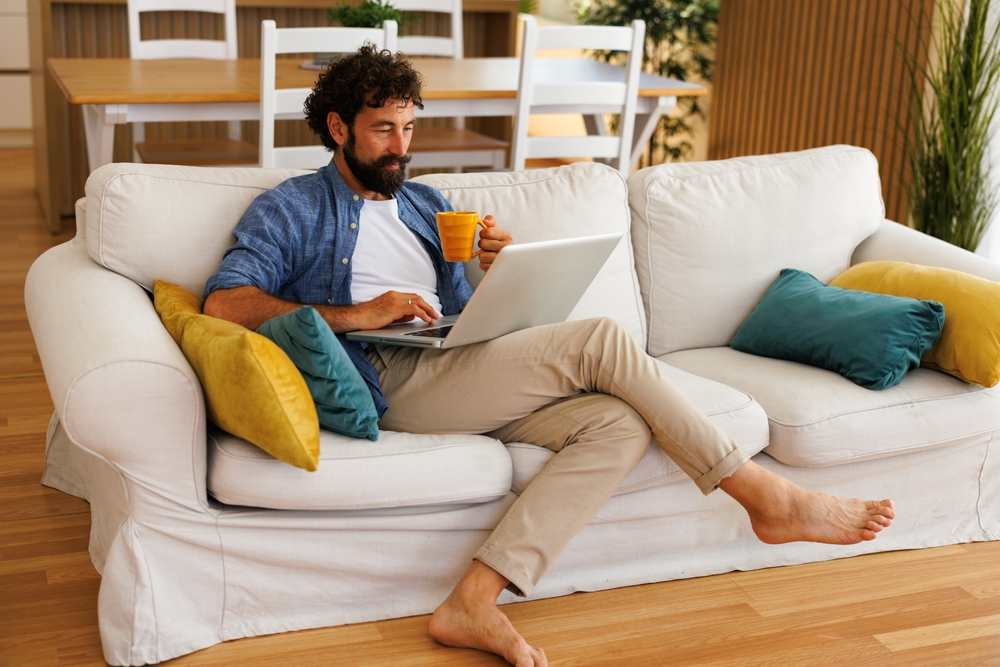 Man at home enjoying coffee and using laptop on sofa