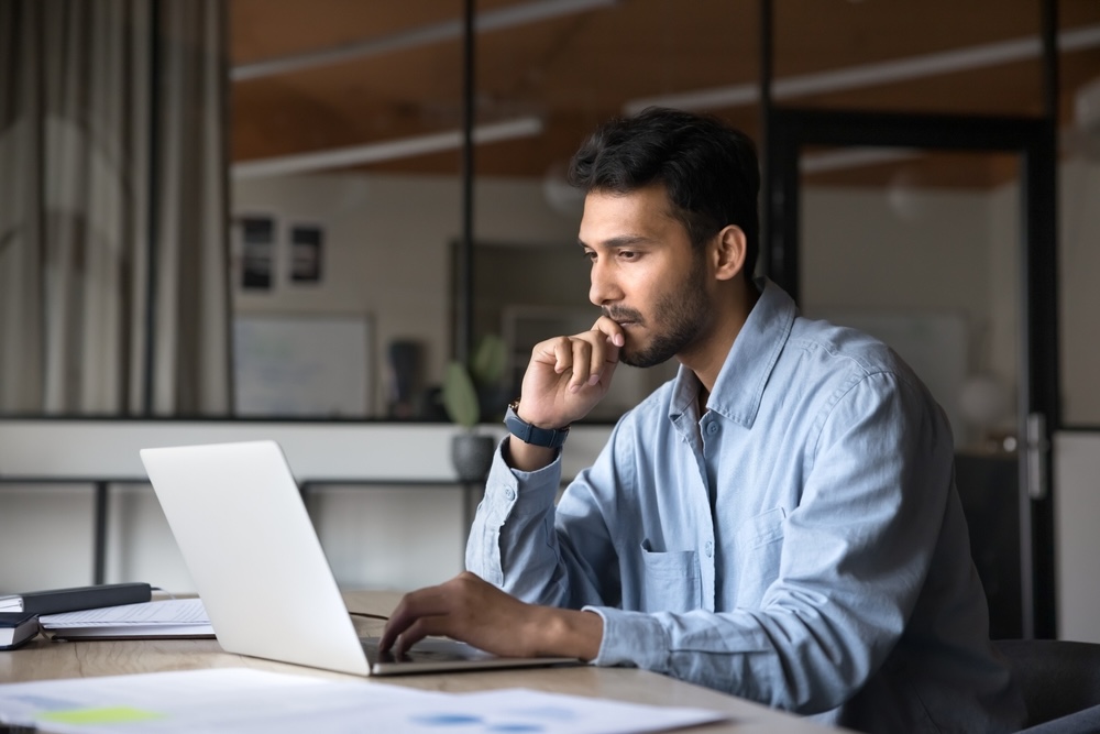 man working at laptop, sitting at workplace table, looking at display, typing, touching chin