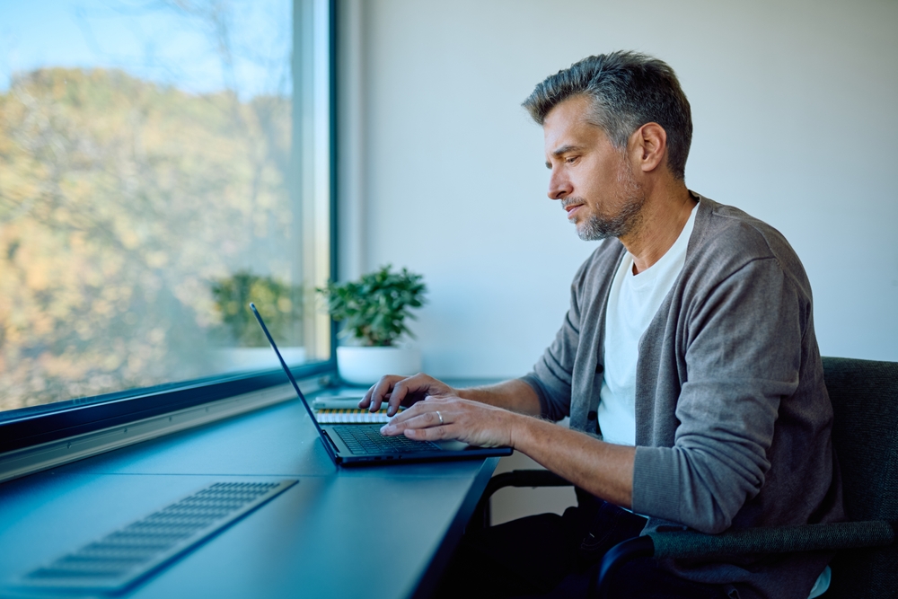Focused man typing on laptop, networking from home office. Professional connection by window