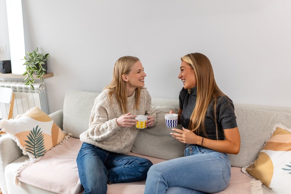 Two young women laughing and drinking coffee on sofa