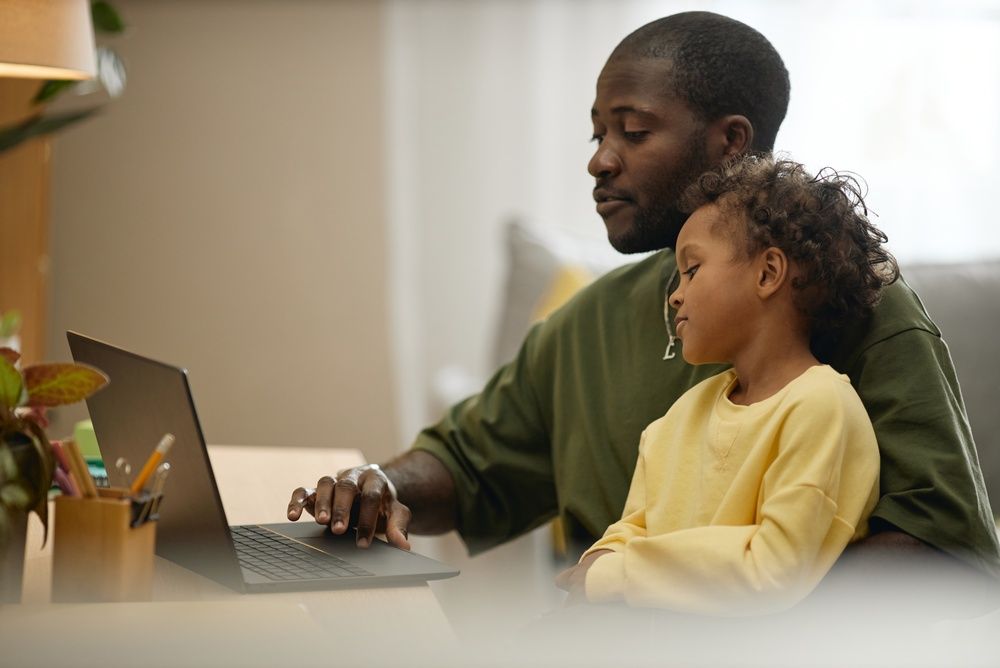 father working on laptop with child son sitting on lap at home desk