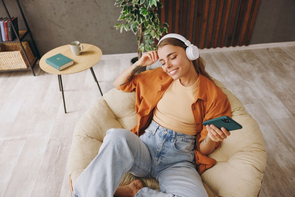 young woman wears casual clothes listen music in headphones use mobile cell phone sits in chair stay at home